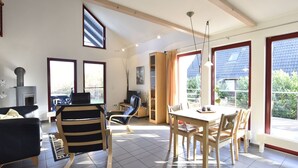 A spacious living room with a fireplace, a dining area with a table and chairs, and large windows offering views of the outdoors. - Holiday House on the Island of Poel (Insel Poel)