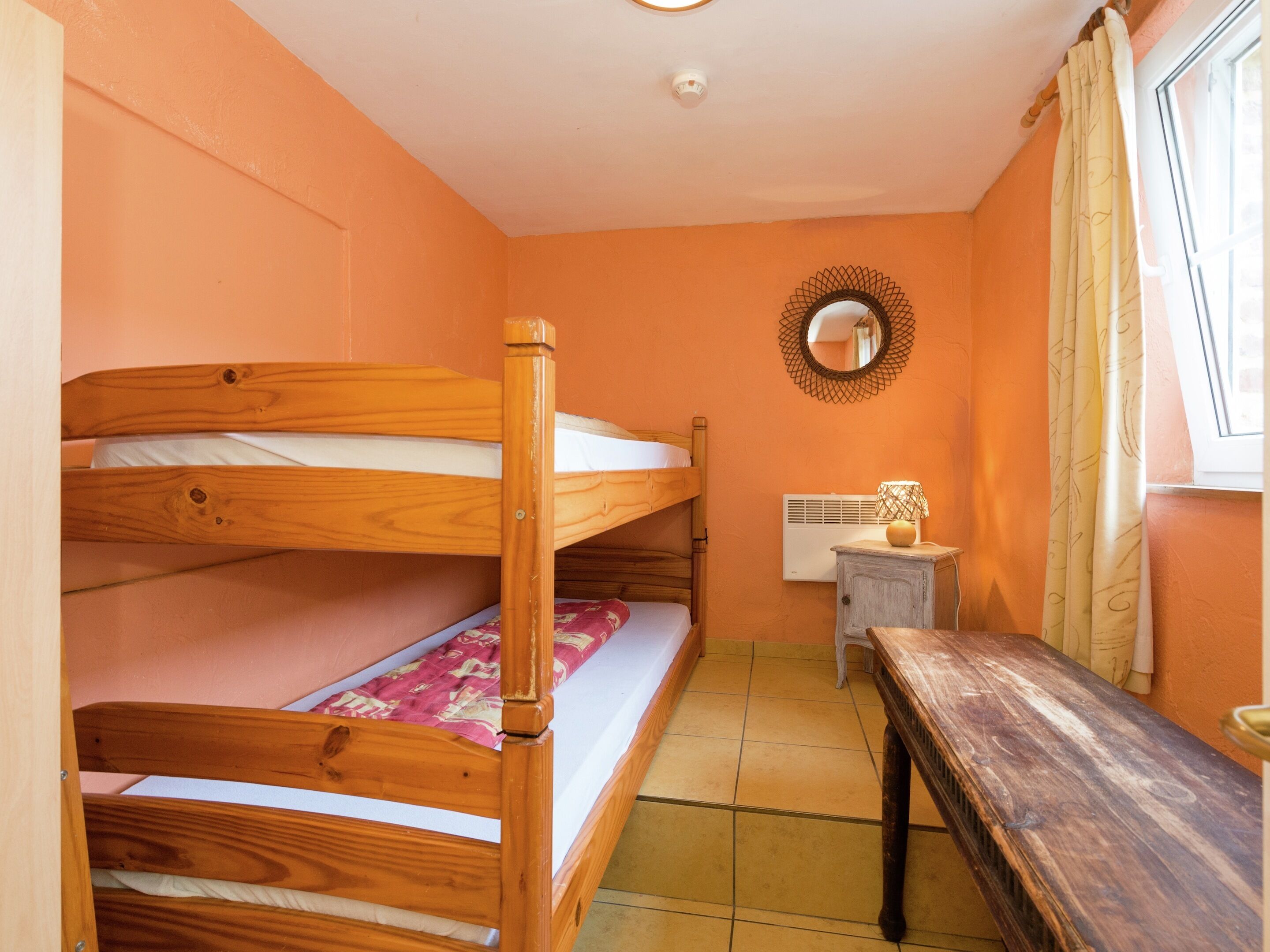 A room with bunk beds, a small table, a mirror, and a window with curtains.
