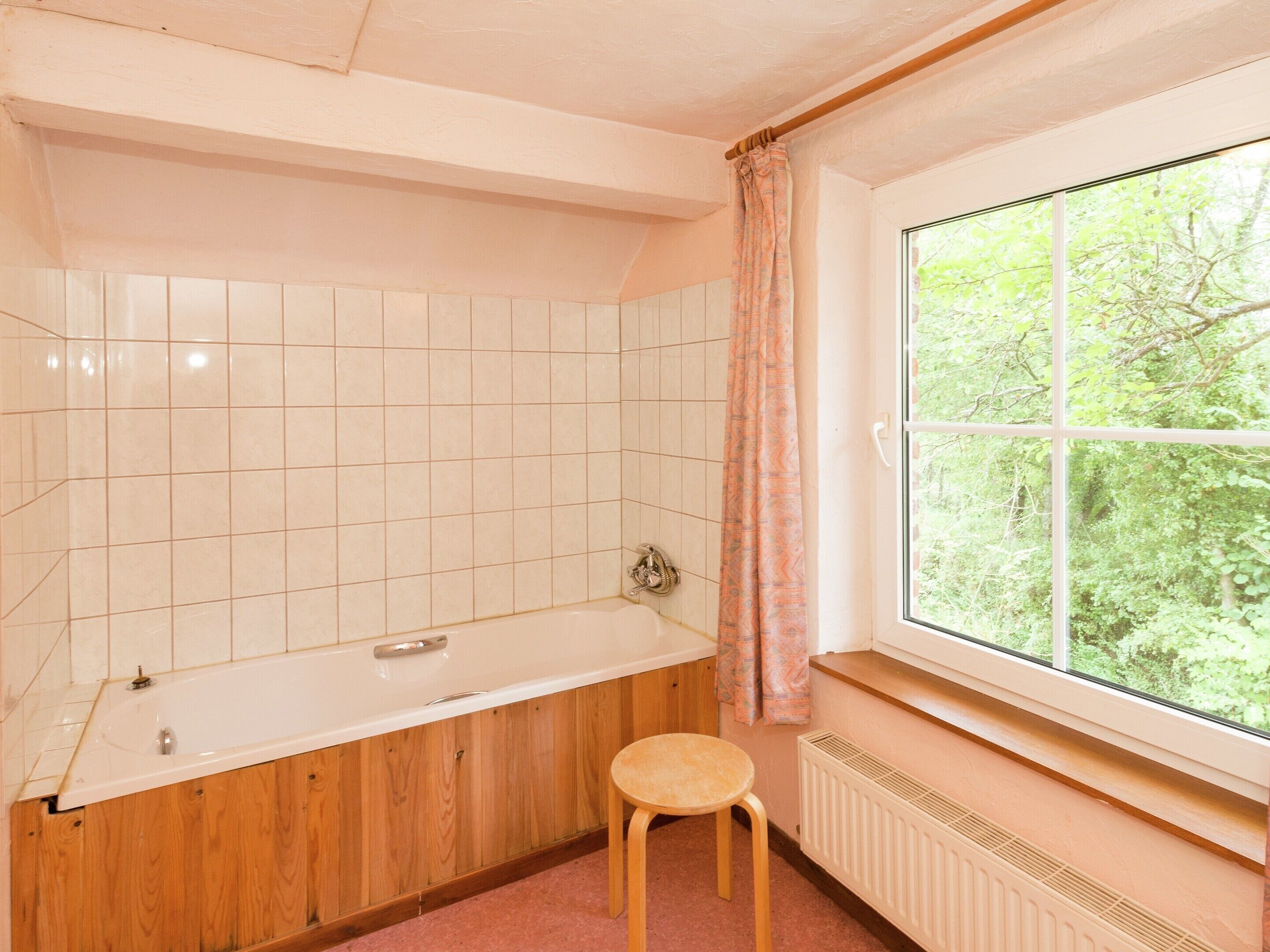 A bathroom with a wooden bathtub, a window with a view of greenery, and a radiator.