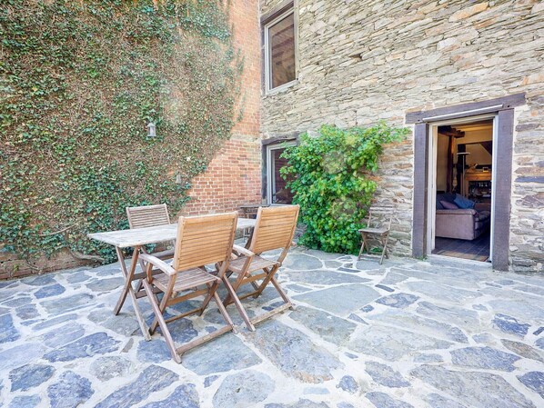 Une terrasse en pierre avec une table et des chaises, un mur de briques recouvert de lierre et une porte d’entrée donnant sur un espace intérieur.