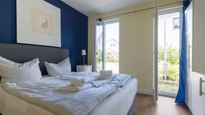 Un lit bien fait, avec du linge de lit blanc et une serviette enroulée, une table de chevet avec une lampe, et une grande fenêtre donnant sur un bâtiment et de la verdure.