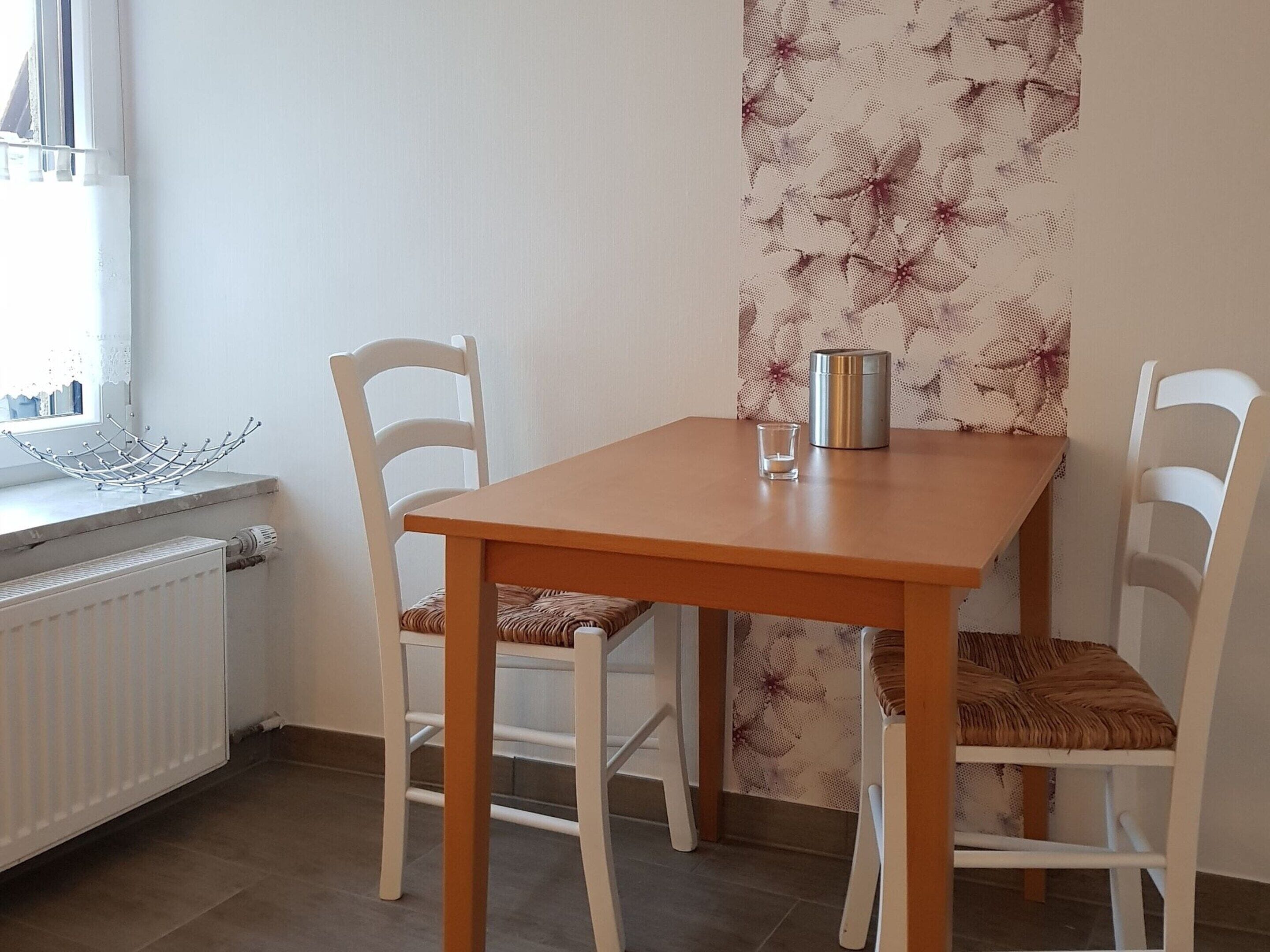 A small dining area with a wooden table and white chairs, a radiator, and a window with a decorative metal piece on the sill.