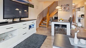 A modern living room with a flat-screen TV, a dining area with a table and chairs, a staircase, and a kitchen area. - Grosszugiges Apartment in Boltenhagen am Meer (Boltenhagen)