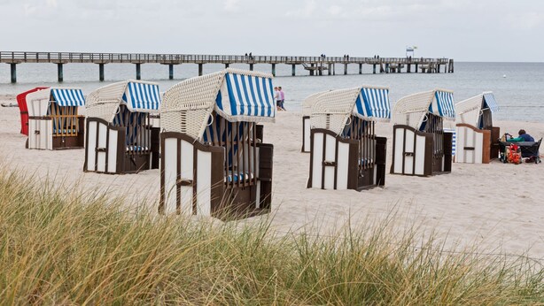 Beach chairs with striped canopies lined up on the sand, a pier in the background, and a person sitting on one of the chairs. - Grosszugiges Apartment in Boltenhagen am Meer (Boltenhagen)