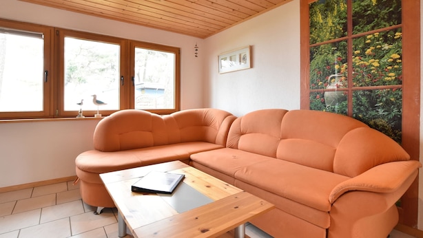 A modern living room with a sectional sofa, a wooden coffee table, and large windows with a view of greenery. - Holiday Home in Am Salzhaff (Am Salzhaff)