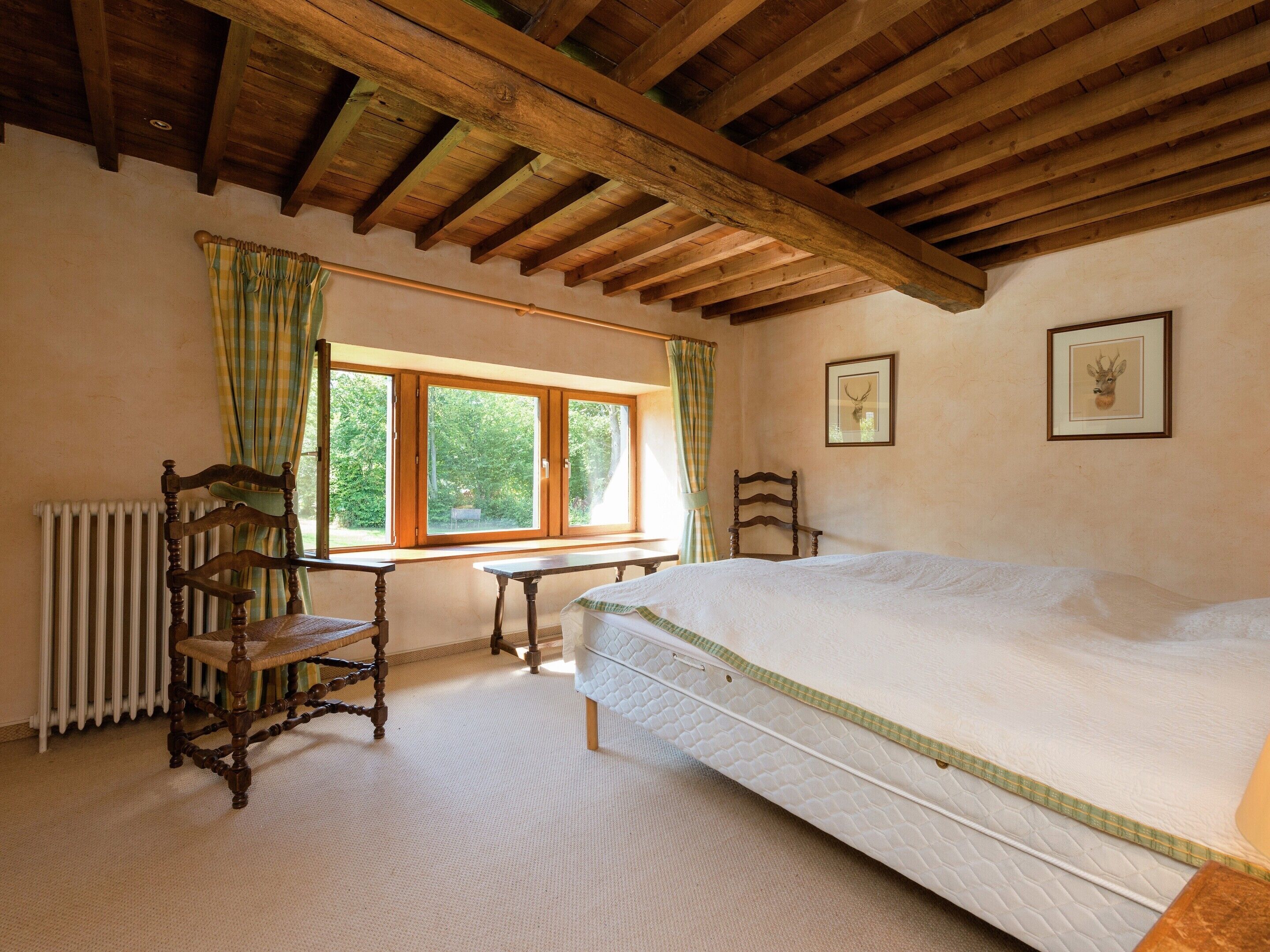 Une chambre avec un grand lit, des poutres apparentes en bois au plafond, une fenêtre avec des rideaux, un radiateur et deux tableaux encadrés accrochés au mur.