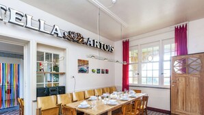 A dining area with a long wooden table set for six, surrounded by wooden chairs. A large sign above the table reads "TELLA ARTOS." The room has a checkered floor pattern, a window with red curtains, and a wooden cabinet to the right.