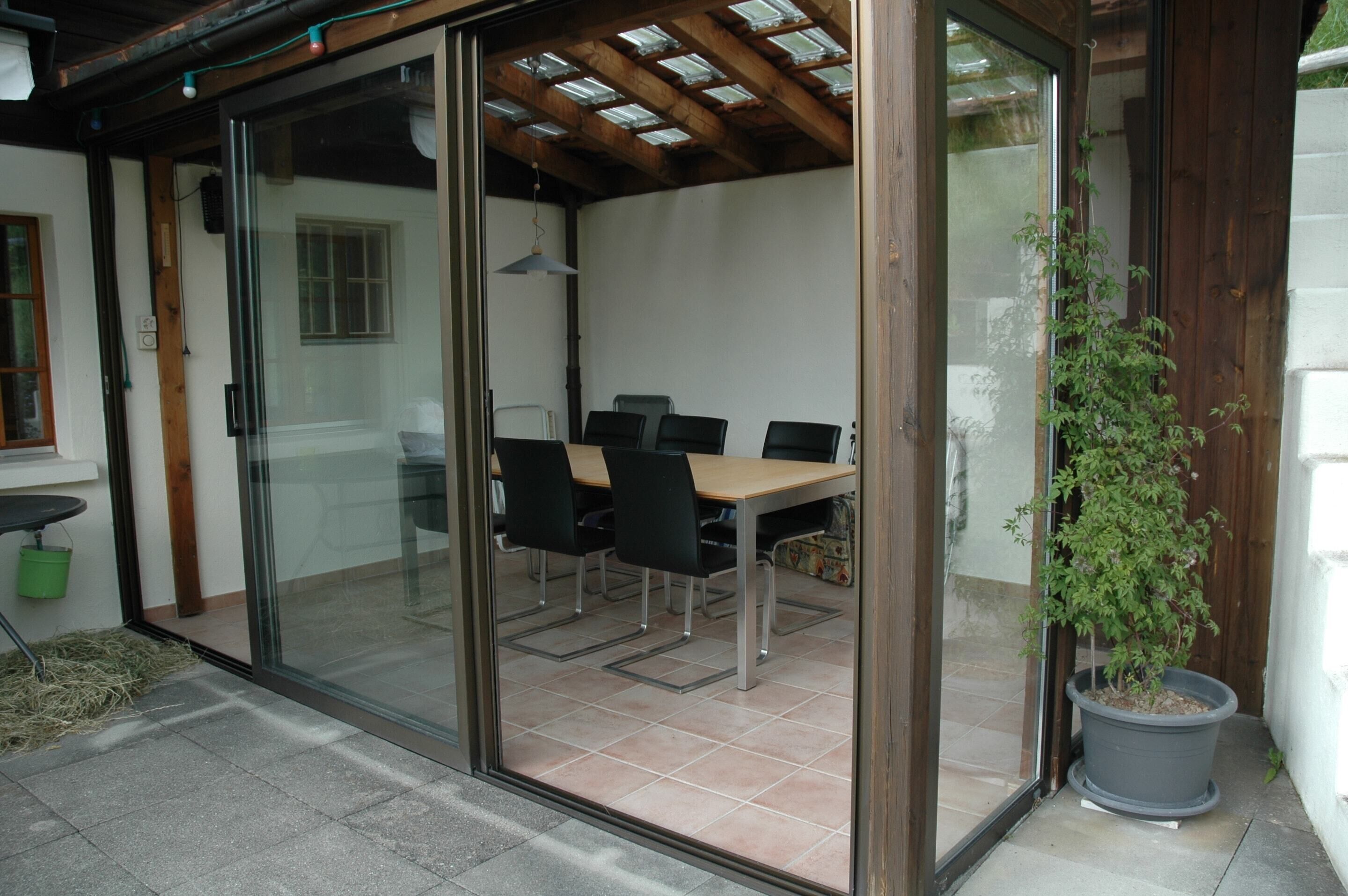 Alpendre coberto com mesa e cadeiras, um vaso de planta e uma porta de vidro deslizante que dá para um espaço interno.