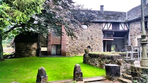 A stone building with a wooden facade, a grassy courtyard, and a stone wall with a metal sculpture. - Apartment in Former Coach House (Waimes)