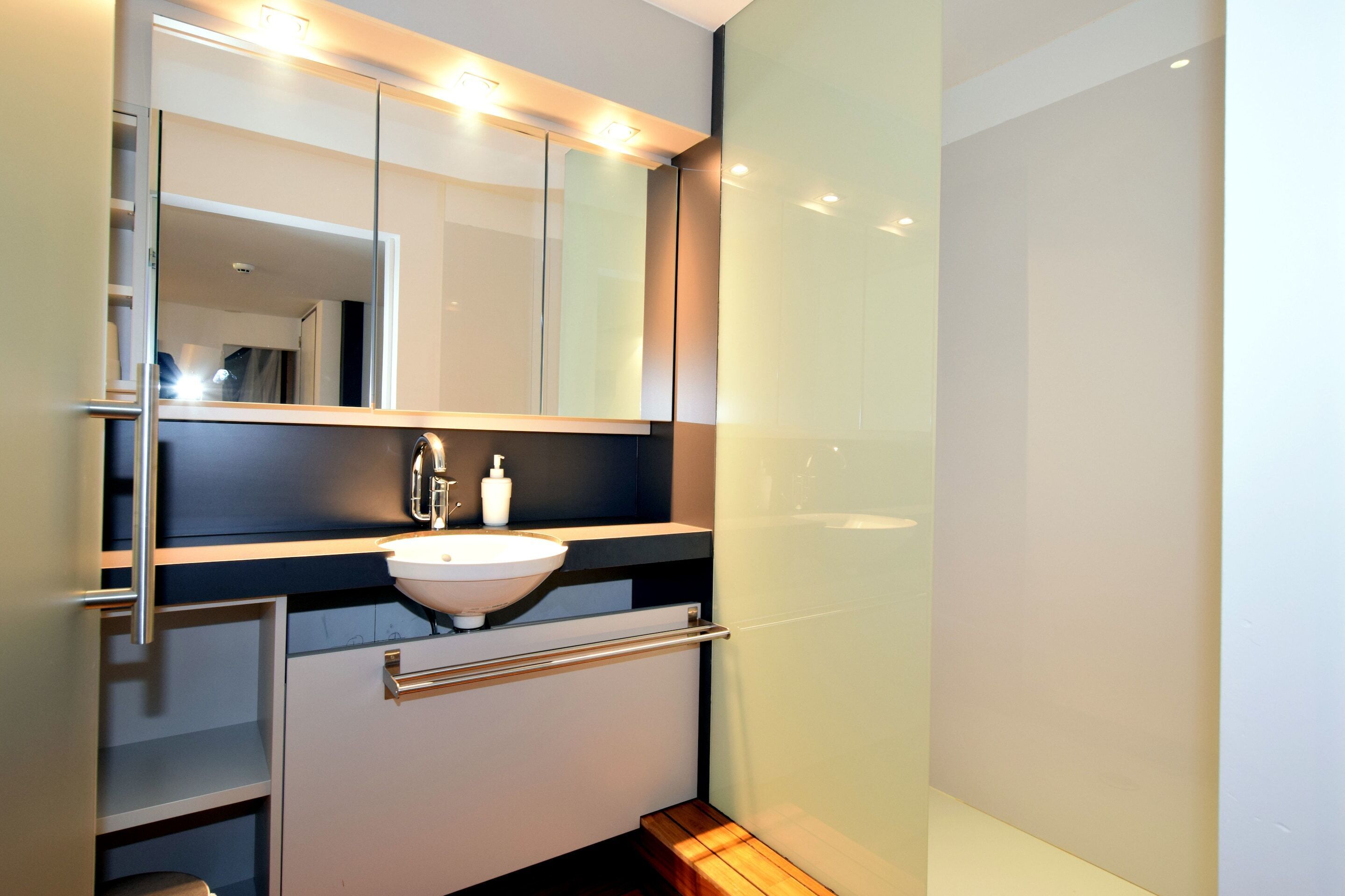 A modern bathroom with a large mirror, a white sink, and a wooden bench.