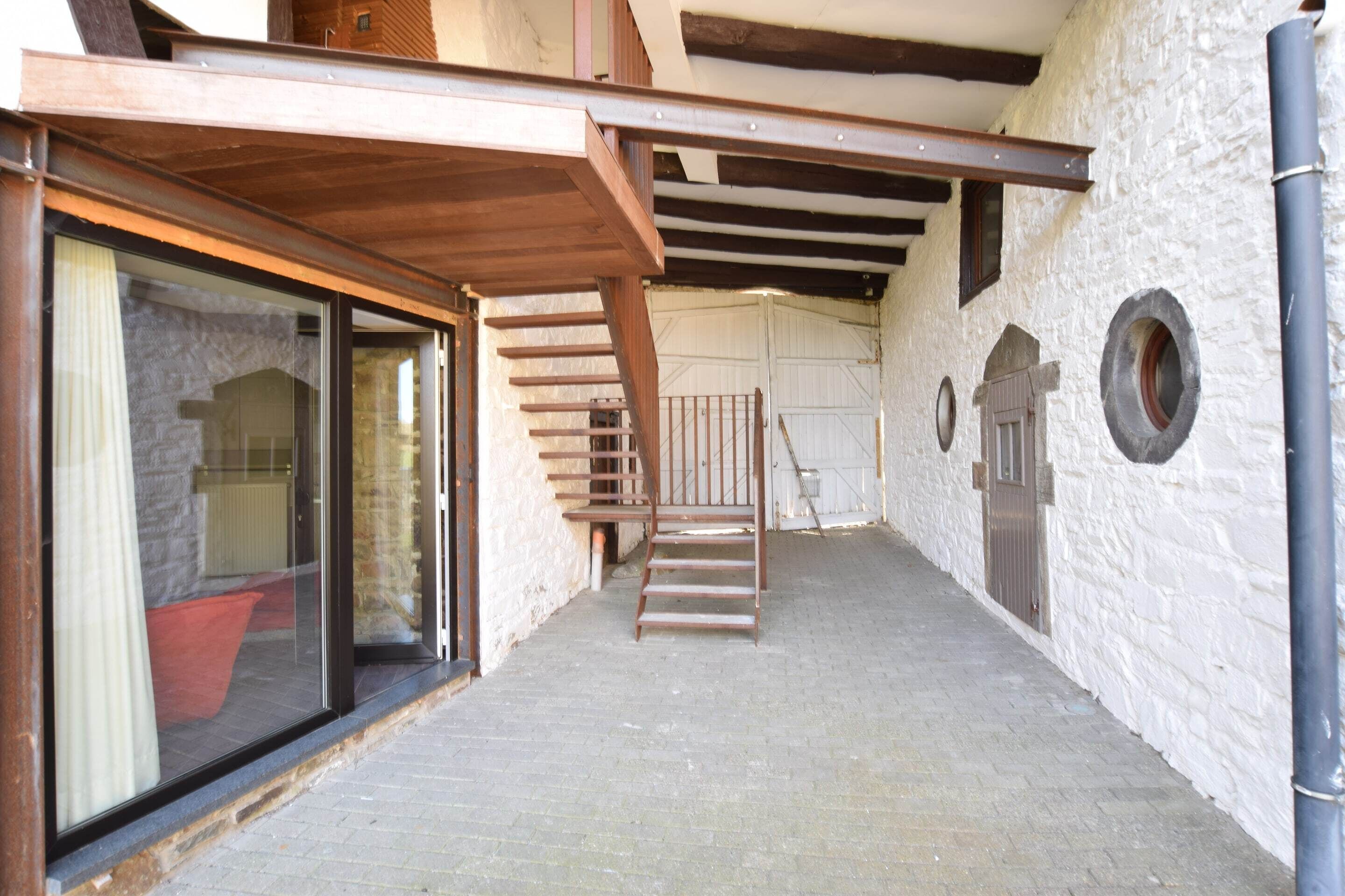 A narrow corridor with a wooden staircase leading to a door. The walls are white and textured, and there is a large window on the left.
