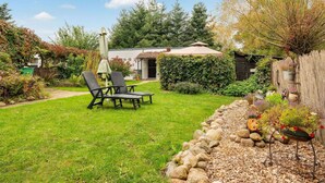 A backyard with a lawn, a wooden fence, a garden bed with plants, and a patio area with a table and chairs. - Quaint Holiday Home near Forest in Stuer (Stuer)