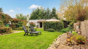 A backyard with a lawn, a wooden fence, a garden bed with plants, and a patio area with a table and chairs.