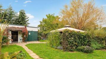A green building with a covered porch, a stone pathway, and a variety of plants and trees.