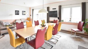 A modern dining area with a long table and chairs, a flat-screen TV mounted on the wall, and a large window with curtains. - Pleasant Holiday Home in Malchow near Beach (Insel Poel)