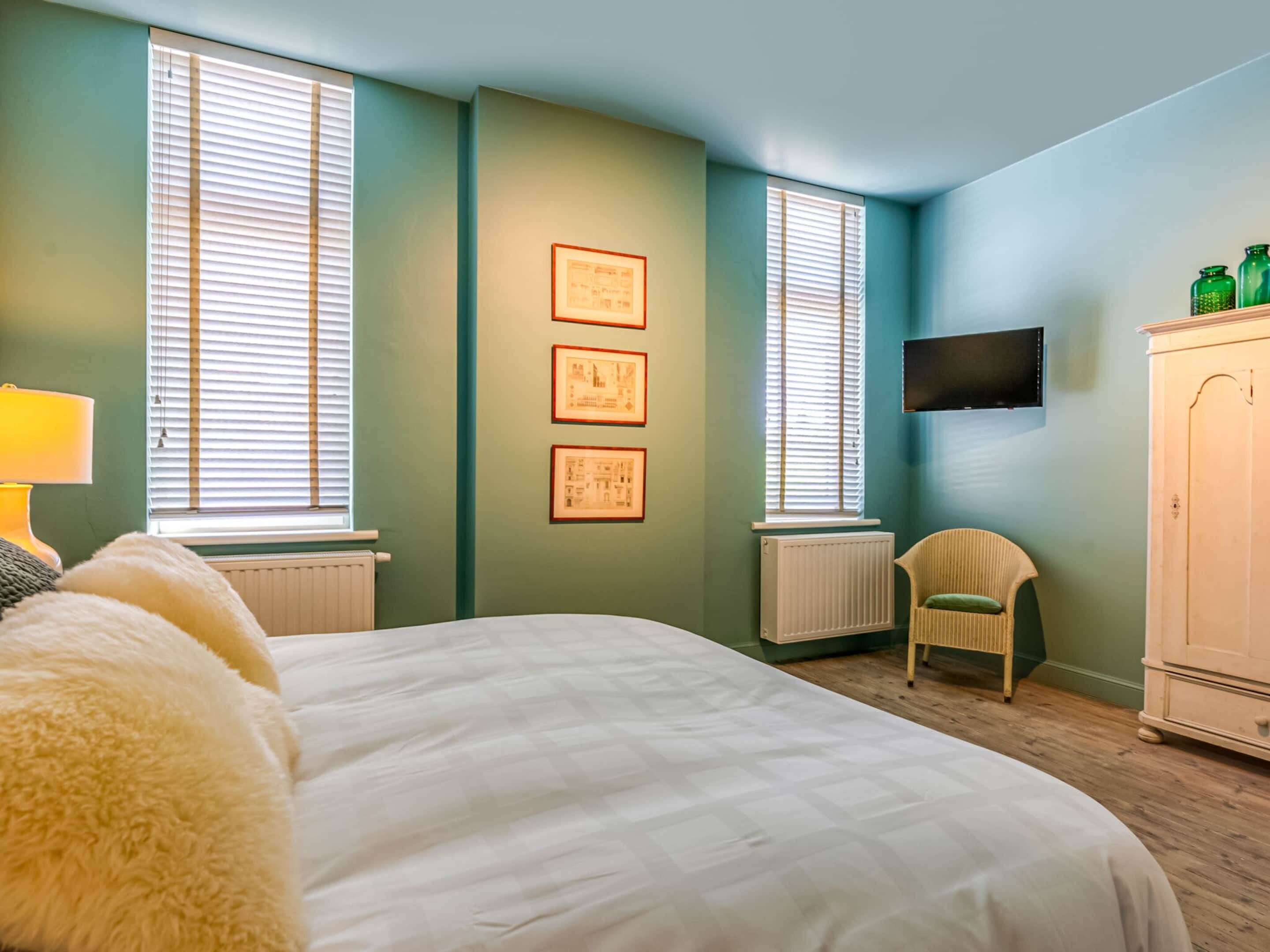 Ein Schlafzimmer mit einem Bett, zwei Fenstern mit Jalousien, einem Heizkörper, einem Sessel, einem an der Wand befestigten Fernseher und einem Schrank.