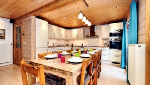 Une cuisine aux murs lambrissés, avec une table à manger pour six personnes, équipée d’appareils électroménagers modernes et d’un luminaire suspendu.
