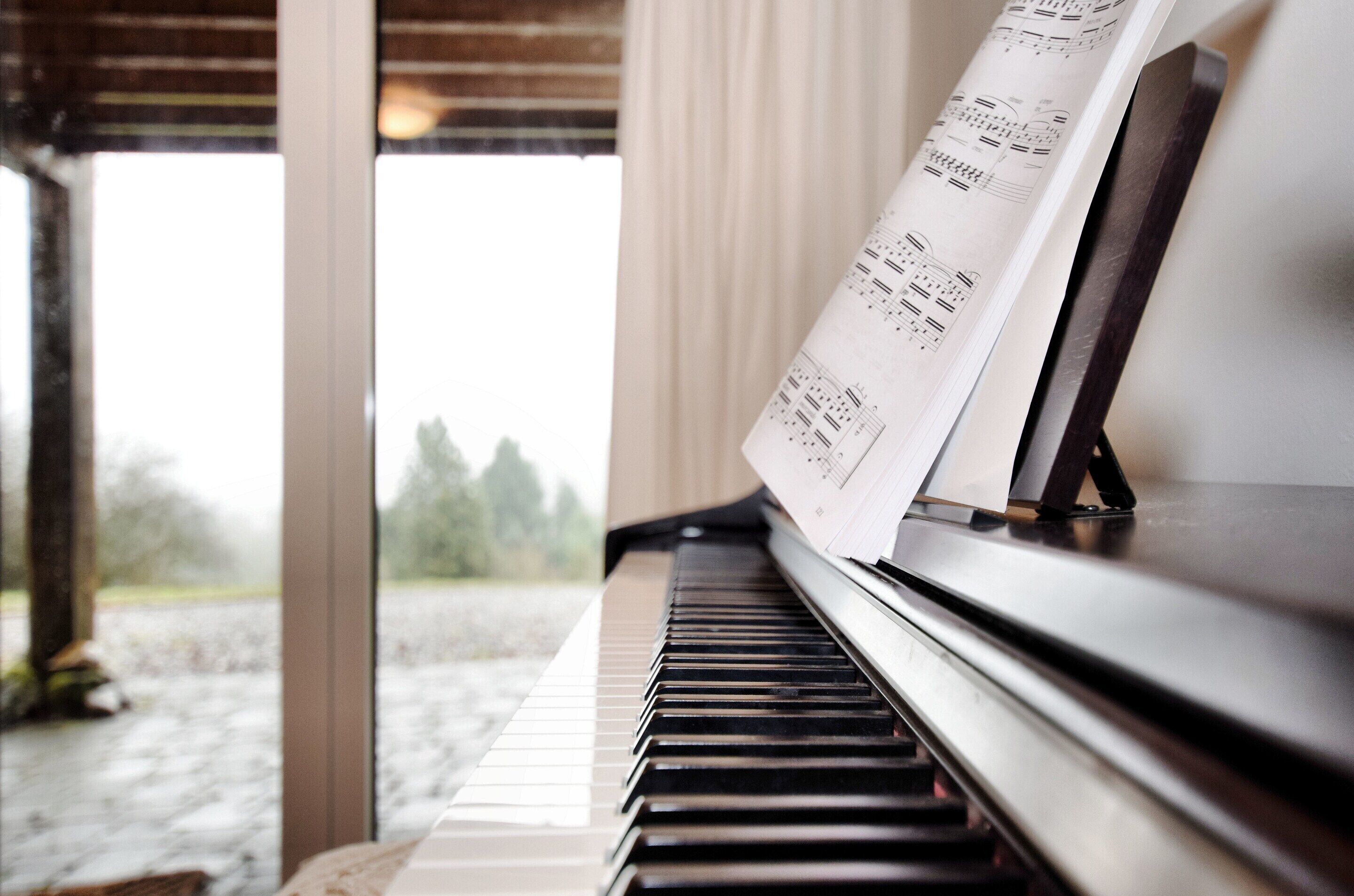 Um piano com partituras em frente a uma janela com vista para árvores e um edifício.