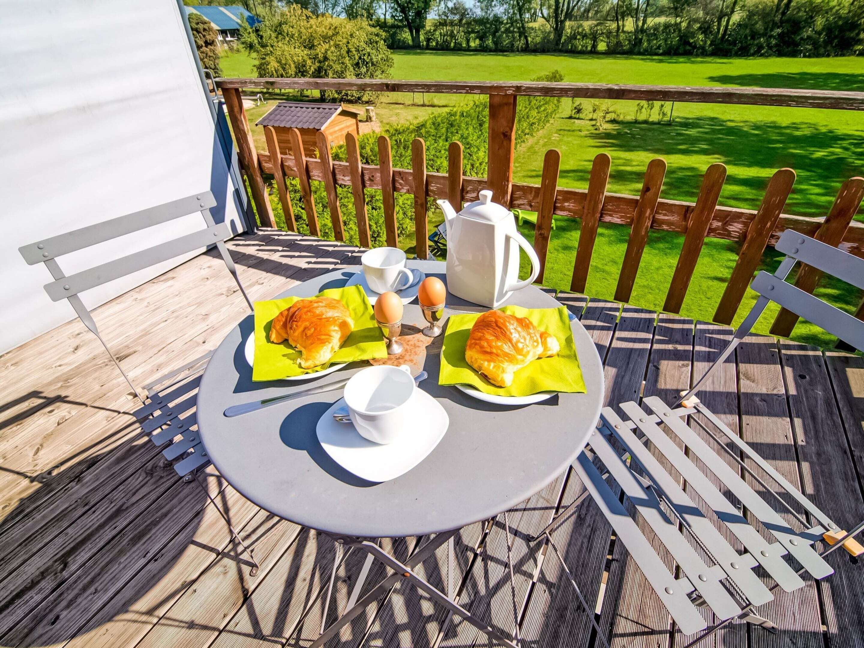 Une table ronde avec un pichet blanc, deux tasses et deux assiettes sur lesquelles sont disposés des croissants, le tout sur une terrasse en bois.