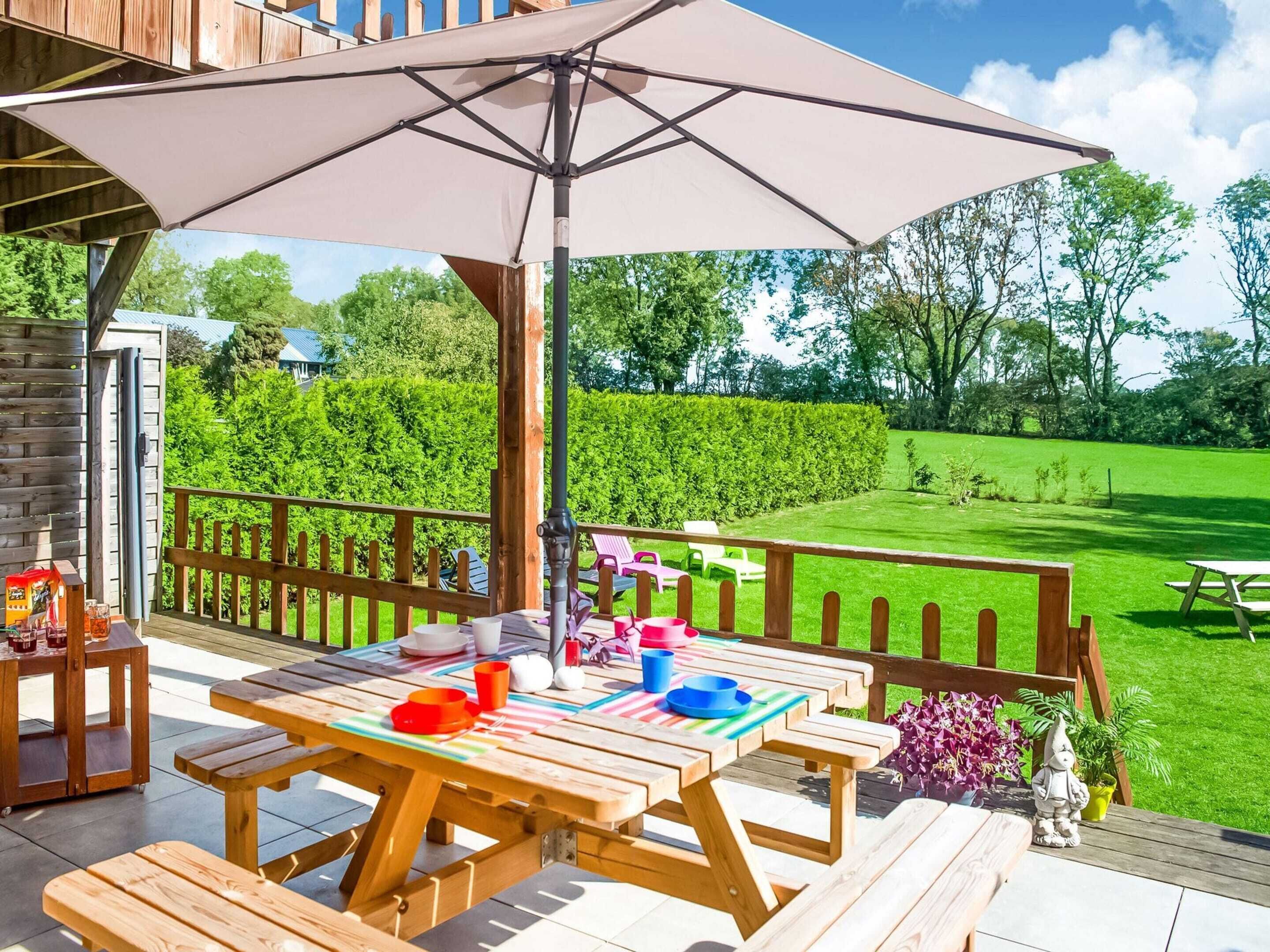 Een houten terras met picknicktafels en een parasol, omringd door een houten hek en een verzorgd gazon.