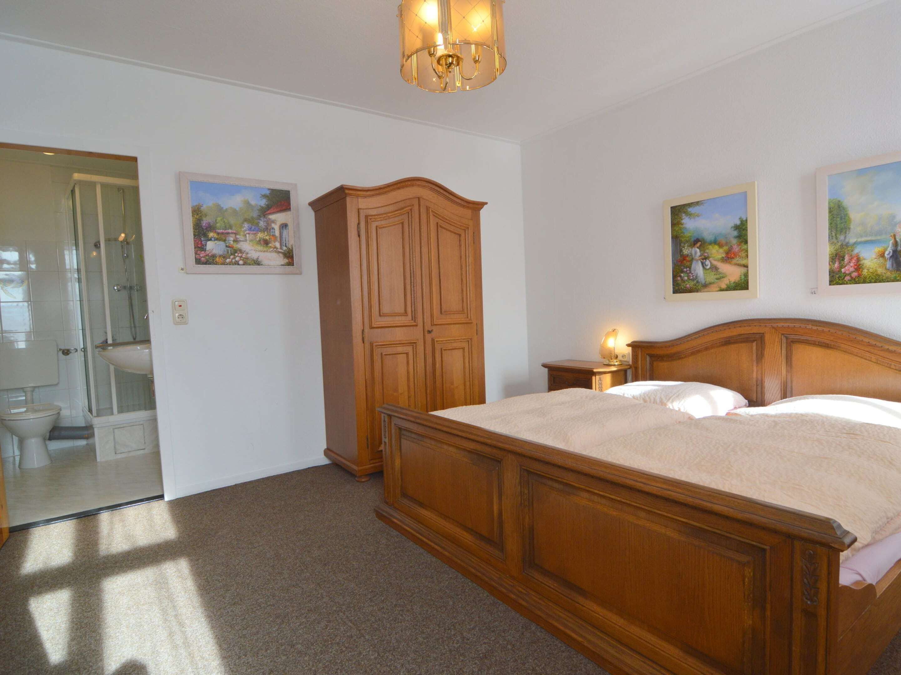 A bedroom with a wooden bed, a nightstand, a wall-mounted lamp, and two framed paintings.