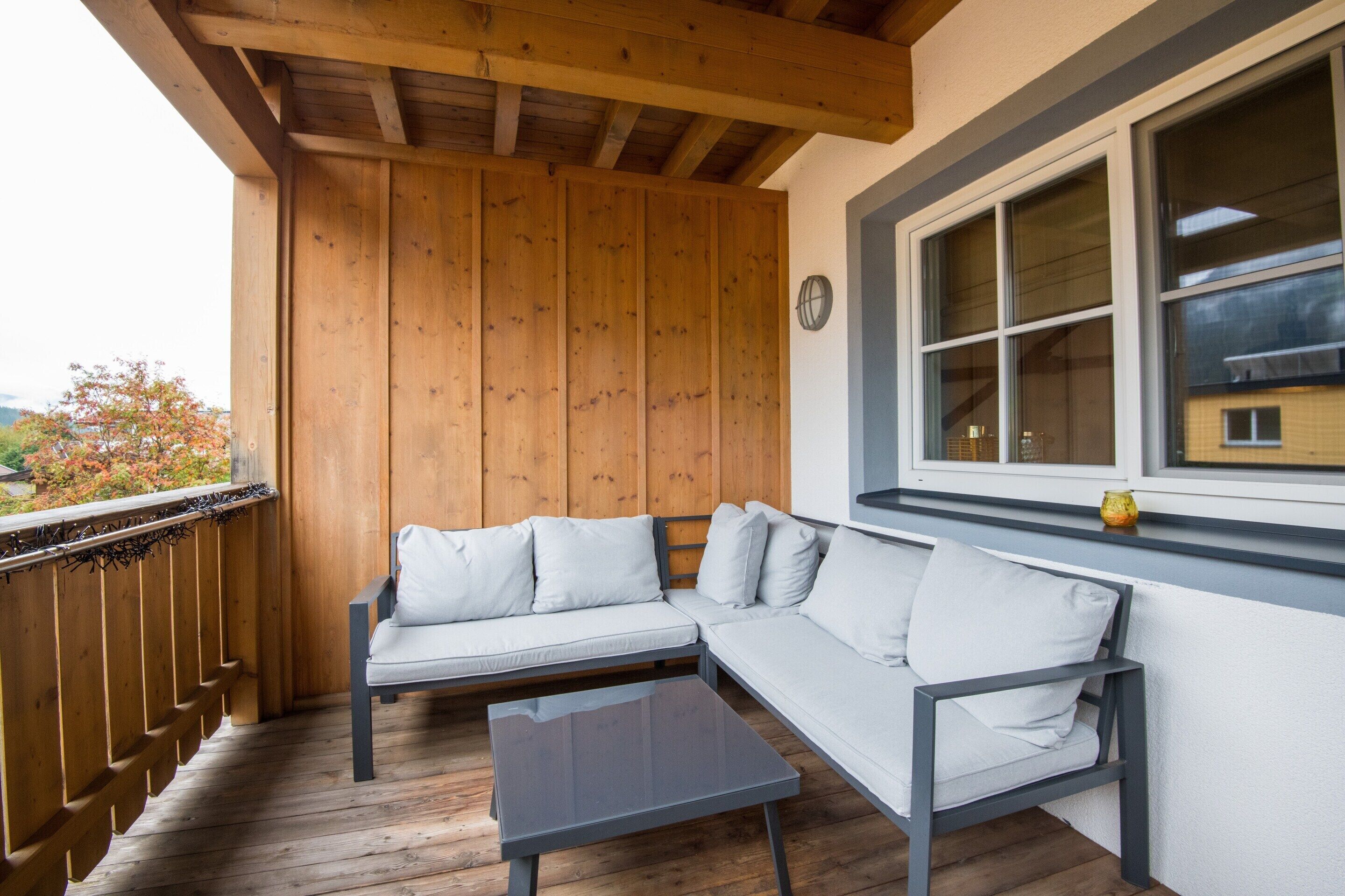 Eine Holzterrasse mit einer Couch und einem Tisch, ein Fenster mit Blick und eine Uhr an der Wand.