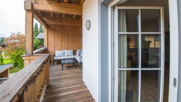Eine Holzterrasse mit einer Couch und einem Tisch, eine Glastür, die ins Innere führt, und Blick ins Freie.