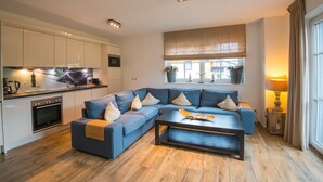 Ein modernes Wohnzimmer mit einer Eckcouch, einem Couchtisch und einer Küchenzeile mit weißen Schränken und einem eingebauten Backofen.