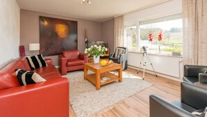 A living room with a red sofa, a wooden coffee table with flowers, a television, and a large window with a view of the countryside. - Ferienhaus in Amelgatzen mit Terrasse (Emmerthal)