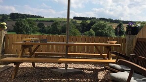 An outdoor seating area with a wooden bench and table, an umbrella, and a view of rolling hills and trees. - Ferienhaus in Amelgatzen mit Terrasse (Emmerthal)