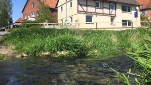 A traditional half-timbered house with a sloped roof, adjacent to a riverbank with overgrown vegetation. - Ferienhaus in Amelgatzen mit Terrasse (Emmerthal)