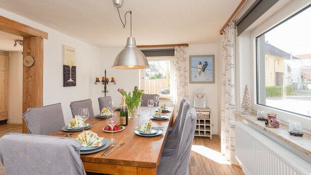 A dining room with a wooden table set for six, grey upholstered chairs, a chandelier, and a window with a view of houses. - Ferienhaus in Amelgatzen mit Terrasse (Emmerthal)