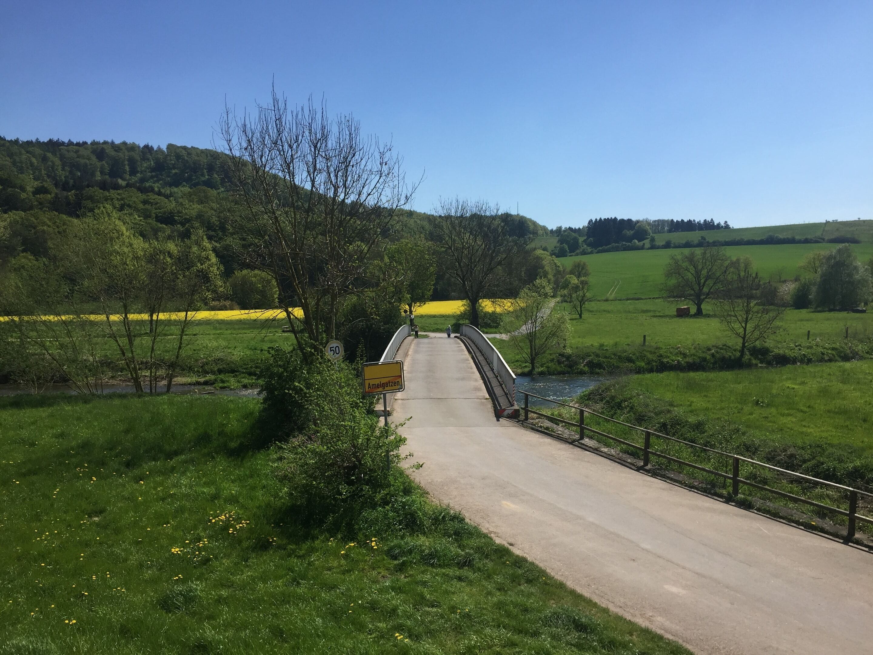 Un camino rural que lleva a un puente sobre un río, rodeado de campos verdes y árboles.