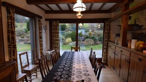A dining area with a long table covered in a black tablecloth with white patterns, surrounded by wooden chairs. A chandelier hangs from the ceiling, and the room has wooden beams. Large windows offer a view of a garden outside. - Hillside Retreat With Pond (Lierneux)