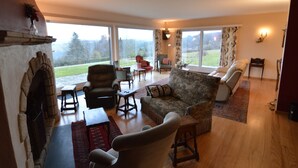 A spacious living room with a fireplace, comfortable seating, a dining area, and large windows offering a view of the outdoors. - Hillside Retreat With Pond (Lierneux)