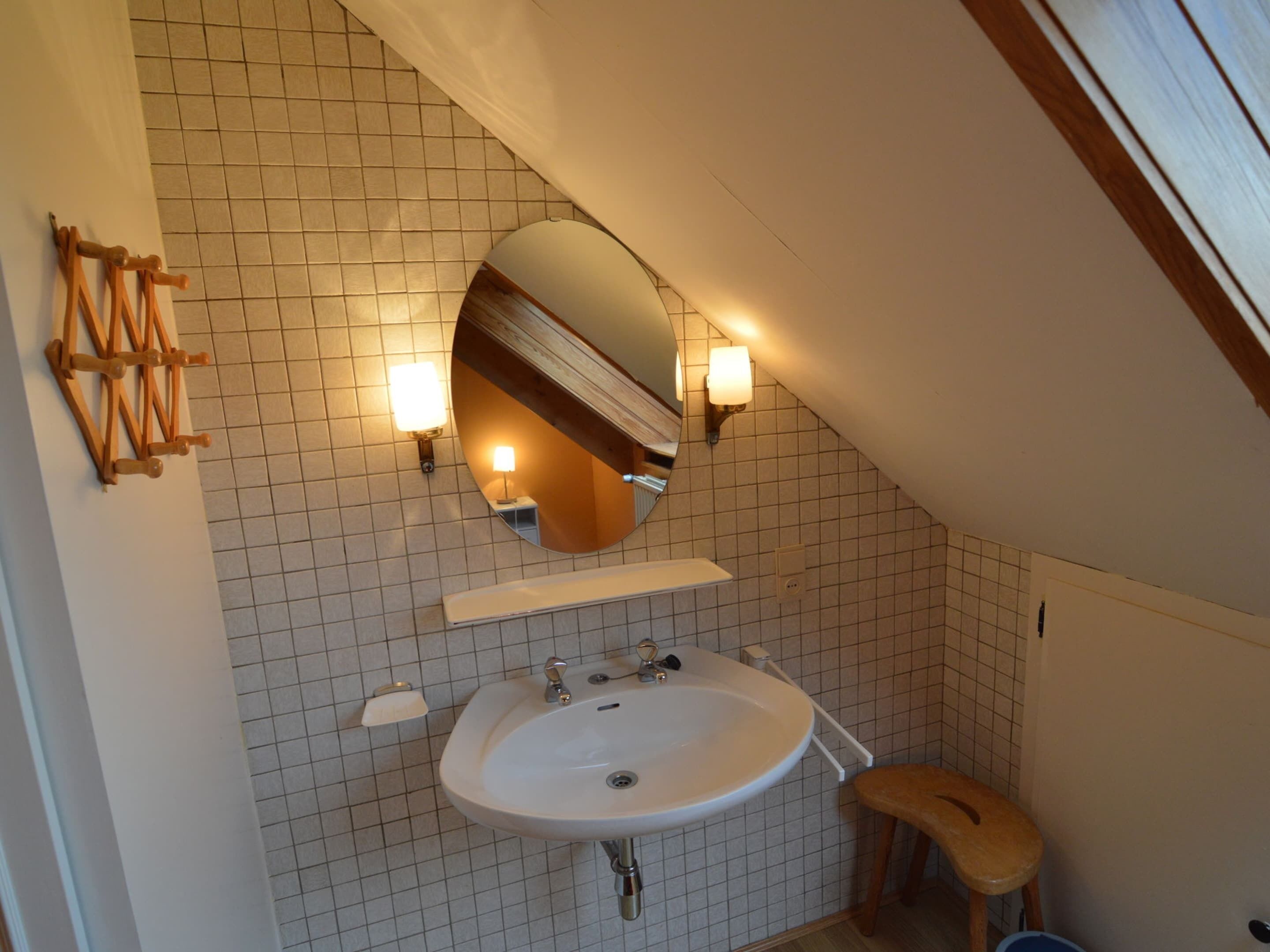 A bathroom with a white tiled floor, a white sink, a wooden stool, and a mirror with two wall-mounted lights.