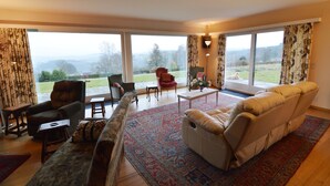 A living room with a sofa, armchair, and a coffee table. There are large windows with curtains, a view of the outdoors, and a lamp on the table. - Hillside Retreat With Pond (Lierneux)