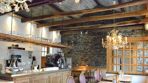 Ein rustikaler Innenraum mit Holzbalken, Steinwänden und einer Chandelüster. Tische und Stühle sind zum Essen angeordnet.