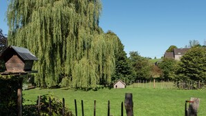 A rural landscape with a weeping willow tree, a wooden birdhouse, a small wooden structure, and a fenced area with purple flowers. - Sprimont Sauna & Garden Stay (Sprimont)