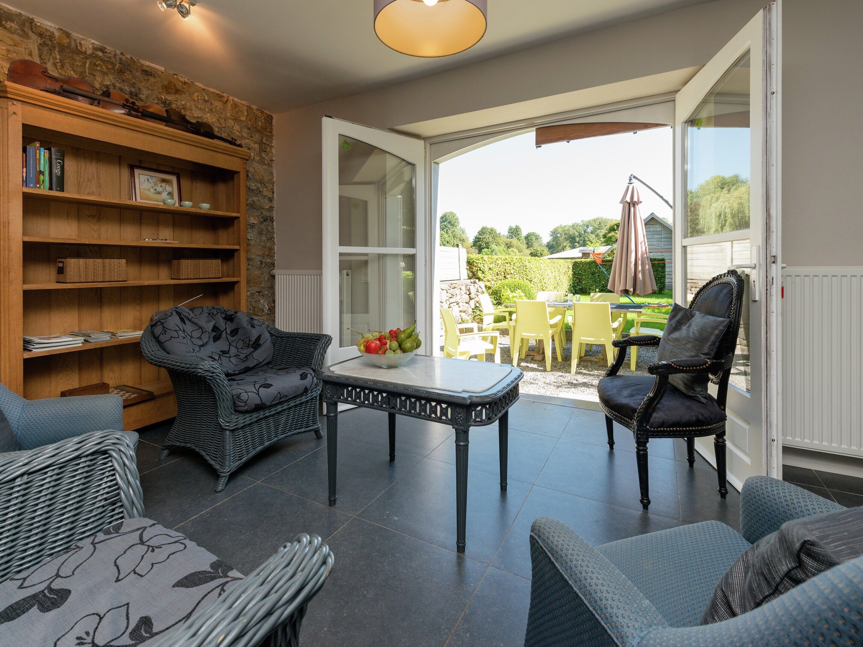 A cozy room with a stone wall, a bookshelf, a wicker sofa, a table with a fruit bowl, and a view of an outdoor patio.