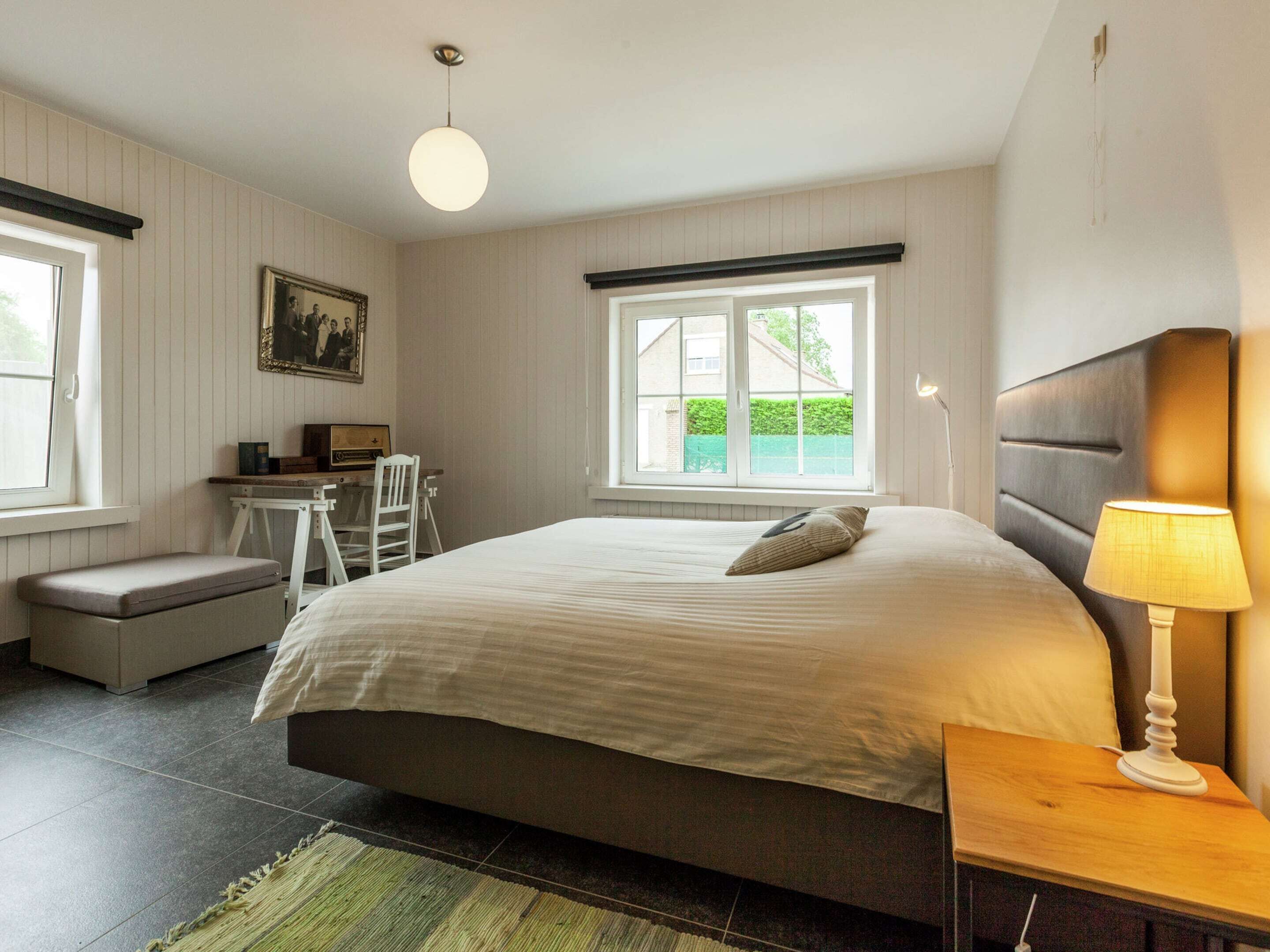 A bedroom with a large bed, a desk, a chair, a bench, and a window with a view of greenery.