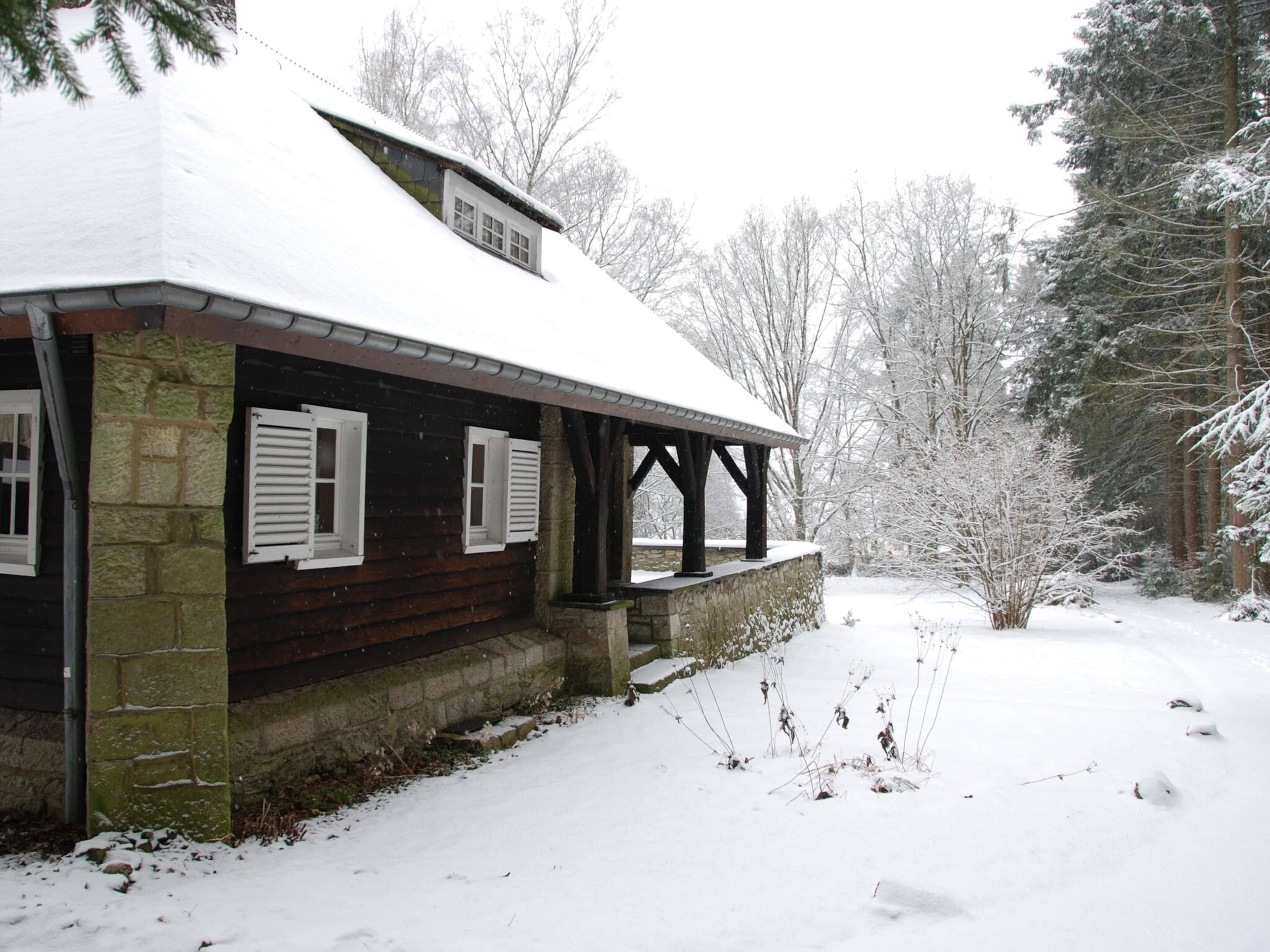 A wooden cabin with a snow-covered roof and stone foundation, surrounded by a snowy landscape and trees.