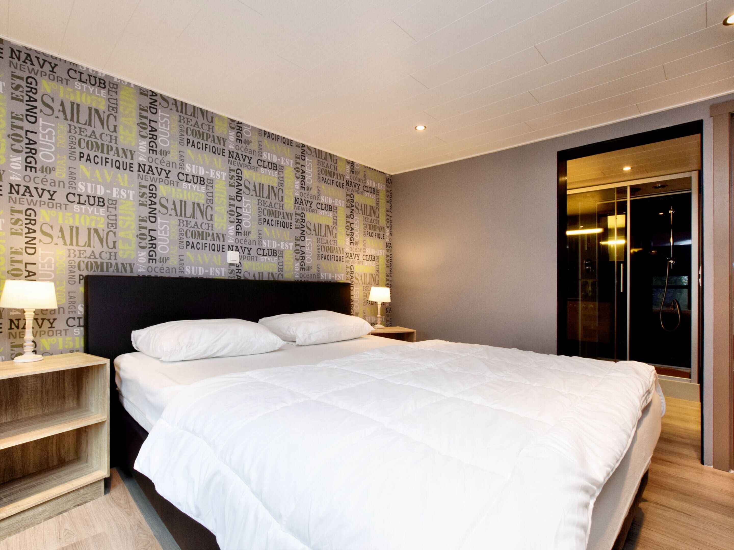 Ein modernes Hotelzimmer mit einem großen Bett, zwei Nachttischen mit Lampen, einer Wand mit maritimem Wandpapier und einer Glastür zum Badezimmer.