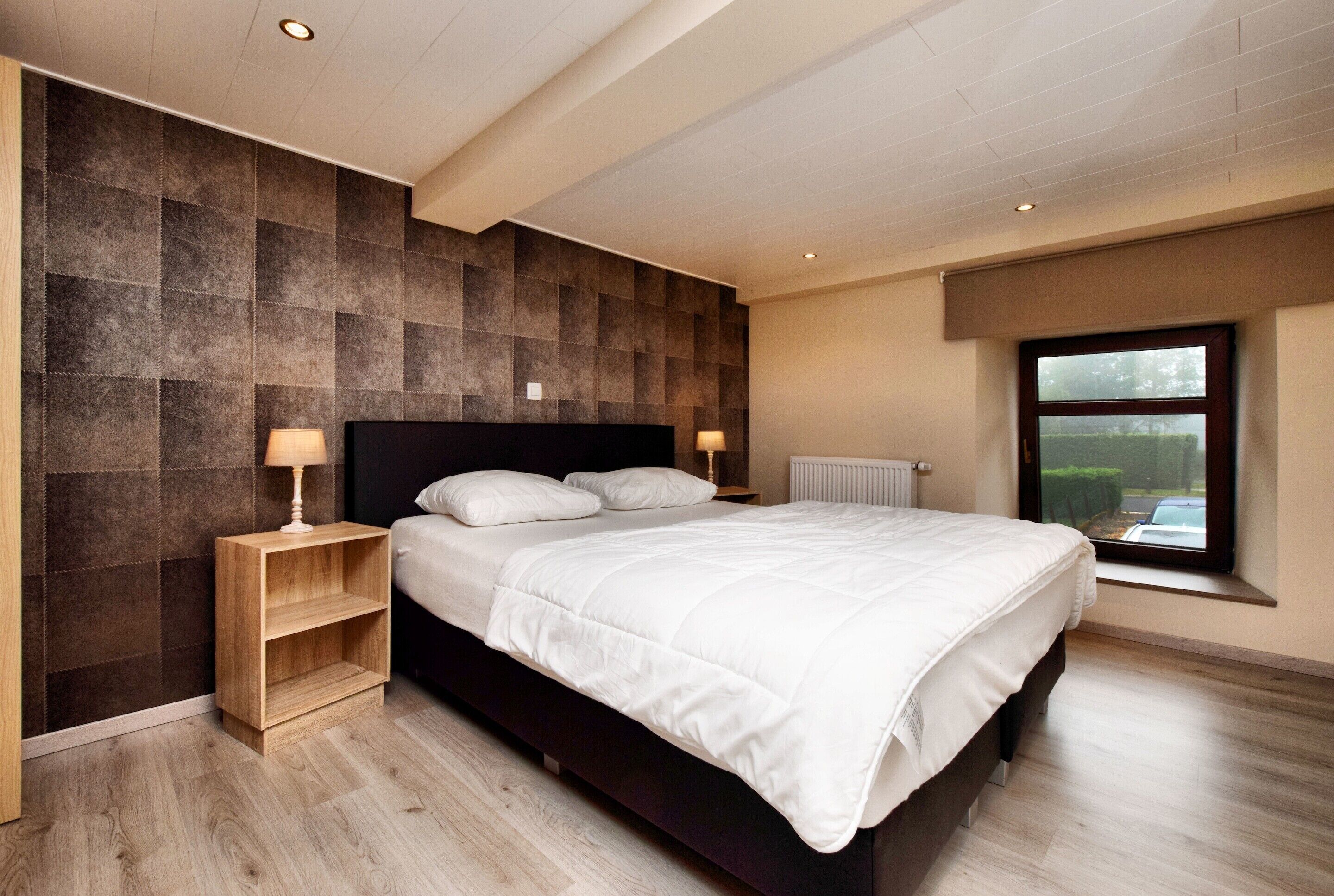 A modern bedroom with a large bed, wooden nightstands, and a view through a window.