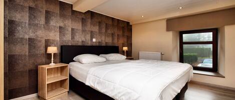 A modern bedroom with a large bed, wooden nightstands, and a view through a window.