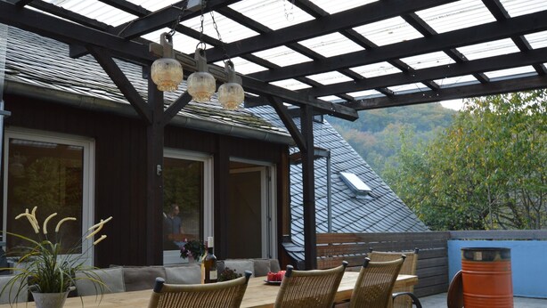 Une terrasse extérieure avec une table en bois et des chaises en osier, un plafond en verre et une vue sur une montagne.