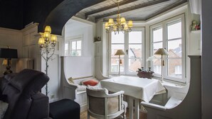 Une pièce chaleureuse avec une fenêtre en saillie, une table recouverte d’une nappe blanche, une chaise et une lampe.