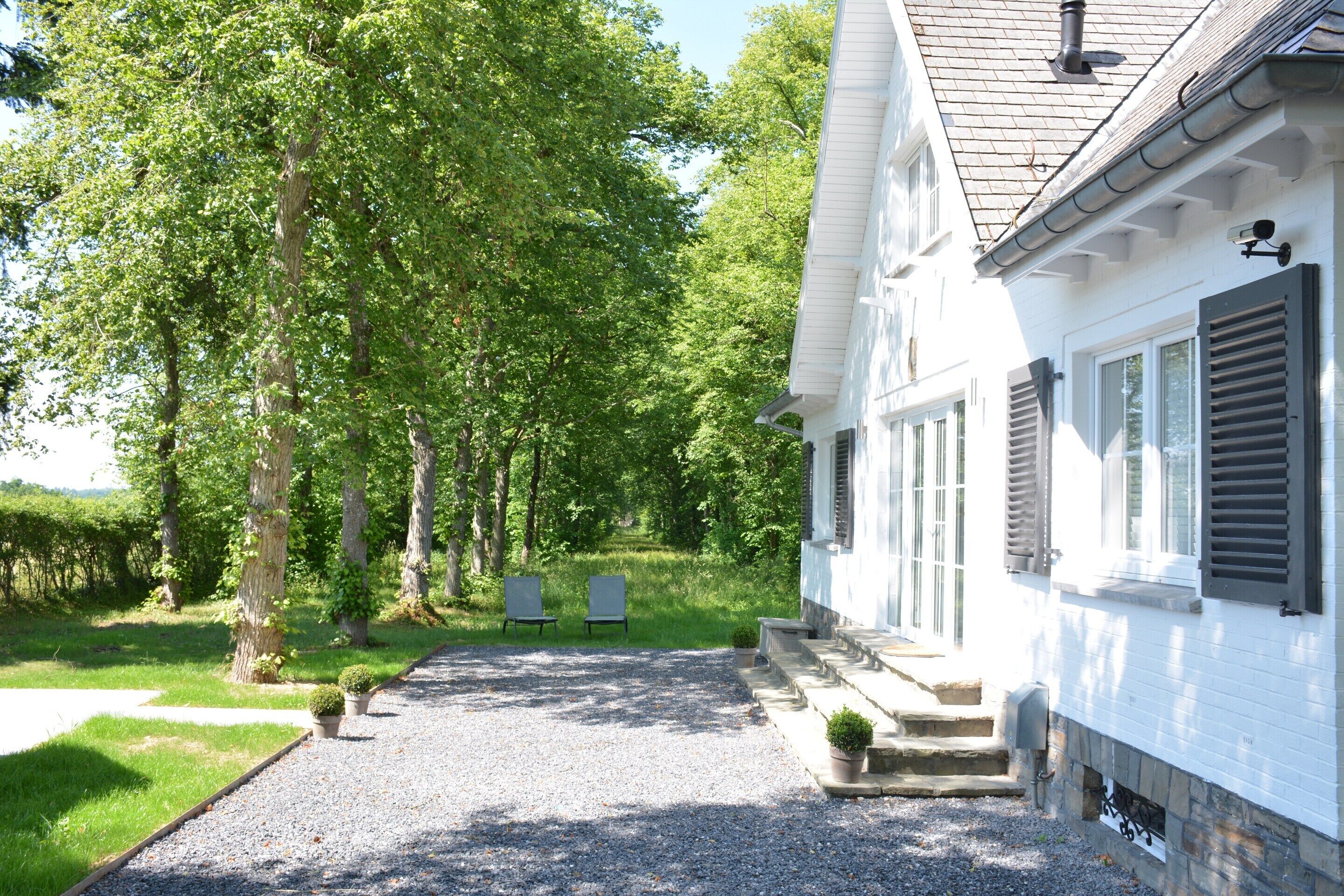 A white building with a gravel driveway leading to a front door. There are two chairs and a small potted plant near the entrance.