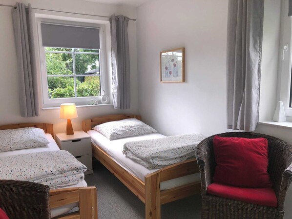 A room with two single beds, a nightstand, a wicker chair, a window with curtains, and a small framed picture on the wall.