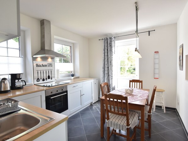A modern kitchen with white cabinets, a built-in oven, and a coffee maker. There is a dining table with chairs and a window with curtains. - Fewo Ostsee Oldenburg/holstein Garten und Carport (Oldenburg in Holstein)